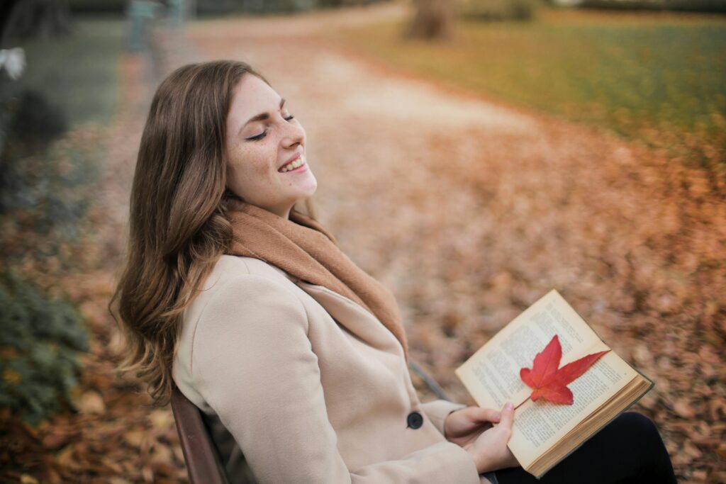 Woman sitting on a bench, smiling while reading a book with a maple leaf in fall.