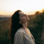 A serene woman with head tilted back, savoring the warm sunset in a field.