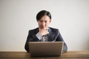 Asian man in suit focusing intently while working on a laptop at a wooden desk indoors.