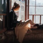 A woman in a leather jacket writes in a notebook while sitting by a sunlit window in a rustic building.