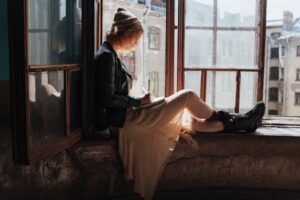 A woman in a leather jacket writes in a notebook while sitting by a sunlit window in a rustic building.