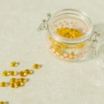 Close-up of open glass jar filled with yellow health capsules on light surface.