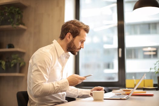 Focused businessman working on laptop while checking smartphone in modern office.