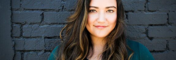 A captivating portrait of a woman smiling confidently in front of a brick wall, showcasing her long brunette hair and vibrant eyes.