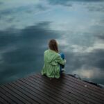 A woman sits on a wooden dock, reflecting by a calm lake under a cloudy sky.