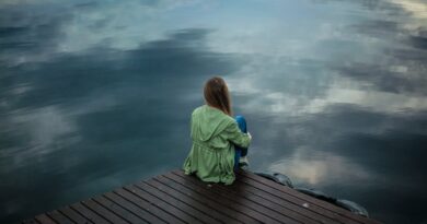 A woman sits on a wooden dock, reflecting by a calm lake under a cloudy sky.