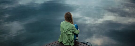 A woman sits on a wooden dock, reflecting by a calm lake under a cloudy sky.