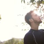 Side view of a young man drinking water from a plastic bottle outdoors in sunlight.