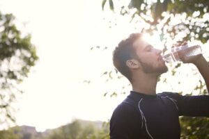 Side view of a young man drinking water from a plastic bottle outdoors in sunlight.