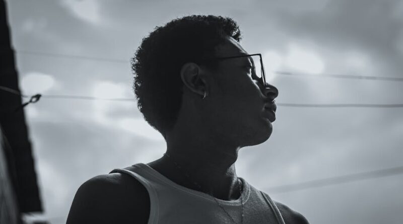 Dramatic side profile silhouette of a reflective young man wearing glasses and a necklace outdoors.