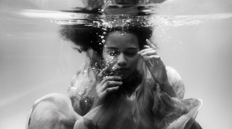 Black and white unemotional young female in loose dress diving with eyes closed into seawater