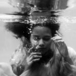 Black and white unemotional young female in loose dress diving with eyes closed into seawater