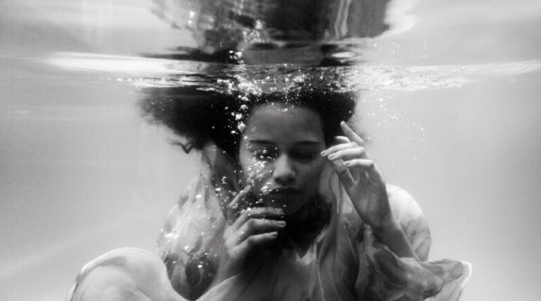 Black and white unemotional young female in loose dress diving with eyes closed into seawater