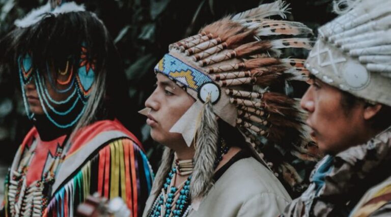 Three Native American men in traditional attire play guitars outdoors, capturing a cultural musical moment.